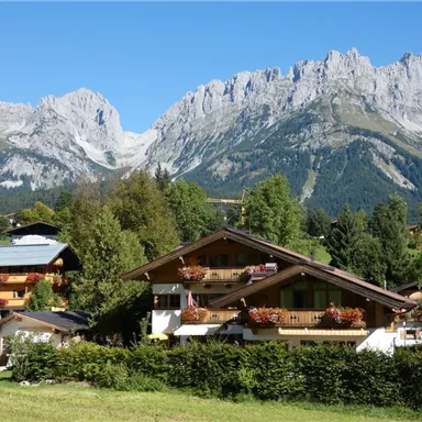 Eine malerische Landschaft mit traditionellen chalets und majestätischen Bergen im Hintergrund. Der Himmel ist klar und blau, was die natürliche Schönheit der Umgebung betont.
