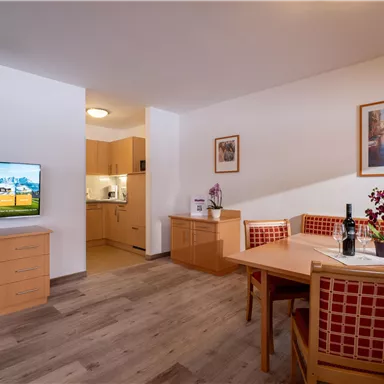 A cozy living area with a dining space and modern furniture. The kitchen is open and well-equipped, with a television on the wall.