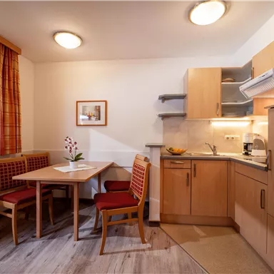A cozy kitchen with a dining area and modern wooden cabinets. The table is decorated with a flower, and the light is warm and inviting.