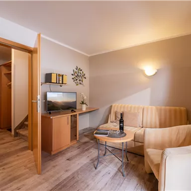 A cozy living room with a sofa, a chair, and a small table. The walls are light, and there is a television as well as warm wood details.