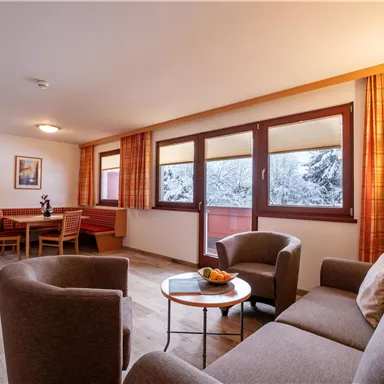 A cozy living room with a sofa, two armchairs, and a table. Large windows show a snowy landscape outside.