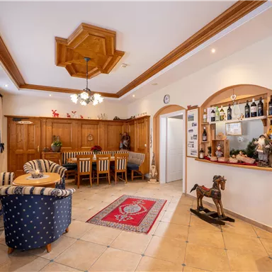 A cozy lobby with wooden furniture and a large dining table. On the counter, drinks and decorative elements can be seen.