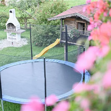 Ein Trampolin mit einem gelben Rutschen liegt in einem grünen Garten. Im Hintergrund ist ein Holzhaus und ein Grill zu sehen.