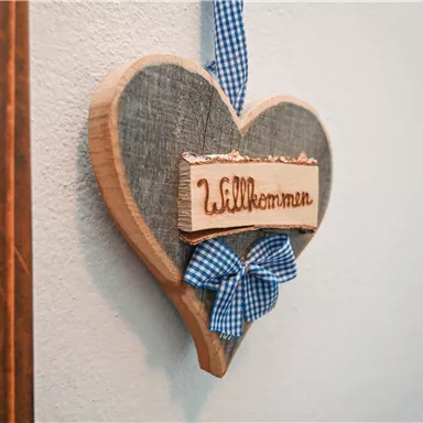 Ein herzförmiges Willkommensschild aus Holz, verziert mit einem blauen Karo-Band. Das Schild hängt an einer Wand und strahlt Gemütlichkeit aus.