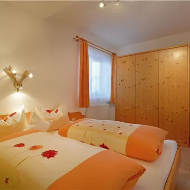 A cozy bedroom with two beds, colorful blankets, and wooden wardrobes. The room is bright with a window and bright light.