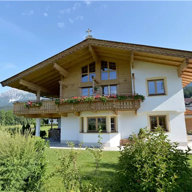 A beautiful, modern house in alpine style with a balcony and flowers. In the background, green meadows and mountains can be seen.