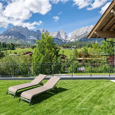 A cozy terrace with loungers on green grass. In the background, impressive mountains and a clear blue sky can be seen.