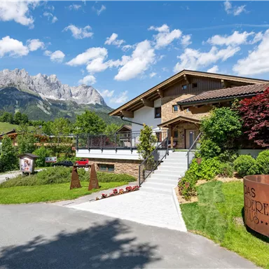 A modern building nestled in a picturesque landscape with mountains in the background. The entrance is surrounded by colorful plants and the sky is clear and blue.