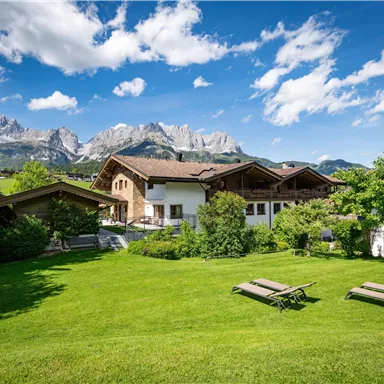 A beautiful landscape with a green meadow and a view of the mountains in the background. The building in the foreground has traditional alpine architecture.