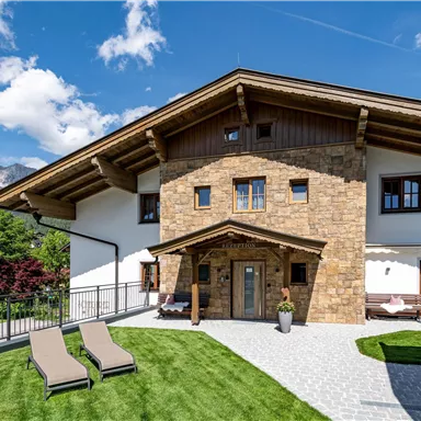 A beautiful house in Alpine style with a well-kept garden. In the foreground, there are loungers, and the mountains are visible in the background.