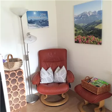 A cozy living area with a brown recliner and a matching footstool. Pictures of the Kitzbühel Alps hang on the wall, and there is a floor lamp as well as decorative baskets.