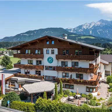 A charming alpine-style hotel with several balconies. In the background, majestic mountains and a green landscape can be seen.