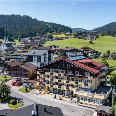 Eine malerische Landschaft mit einem kleinen Dorf in den Bergen. Im Vordergrund steht ein modernes Hotel und im Hintergrund sind grüne Hügel und Wälder zu sehen.