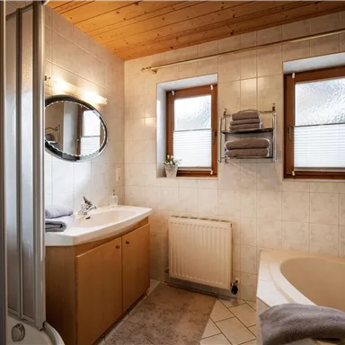 Ein modernes Badezimmer mit Holzdecke und hellen Fliesen. Es gibt eine Badewanne, ein Waschbecken und Fenster mit Plissee.