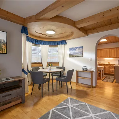 Ein gemütlicher Wohnbereich mit einem Essplatz, Stühlen und einem Holztisch. Die Küche im Hintergrund ist gut ausgestattet und es gibt natürliche Holzdetails im Raum.