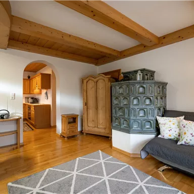 Ein gemütlicher Wohnraum mit Holzdecken und einem traditionellen Ofen. Der Raum ist hell und einladend, mit einem grauen Teppich und bunten Kissen auf dem Sofa.