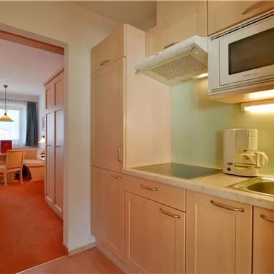 Eine helle Küche mit modernen Geräten und hellen Holzschränken. Im Hintergrund ist ein gemütlicher Wohnbereich mit Tisch und Stühlen sichtbar.