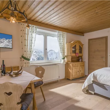 Ein gemütliches Zimmer mit Holzdecke und modernem Fernseher. Es gibt einen Esstisch mit Stühlen, eine gemütliche Bettcouch und große Fenster mit Bergblick.