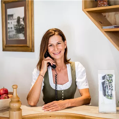 A woman in traditional clothing smiles at the reception and speaks on the phone. Apples and some documents are on the table.