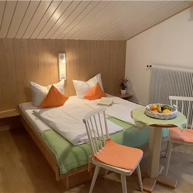 Ein gemütliches Schlafzimmer mit einem Doppelbett und orangefarbenen Kissen. Neben dem Bett steht ein Tisch mit zwei Stühlen und einer Schale Obst.
