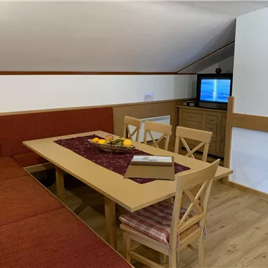 Eine gemütliche Sitzecke mit einem großen Tisch und Stühlen. Im Hintergrund steht ein Fernseher und es gibt eine ansprechende Dekoration.