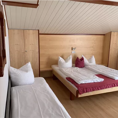 Ein gemütliches Zimmer mit zwei Betten und einem zusätzlichen Bett. Die Wände sind hell, und die Einrichtung ist aus Holz, was eine warme Atmosphäre schafft.