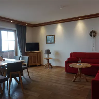 Ein gemütliches Wohnzimmer mit einer roten Couch und einem Holztisch. Große Fenster lassen viel Licht herein und schaffen eine einladende Atmosphäre.