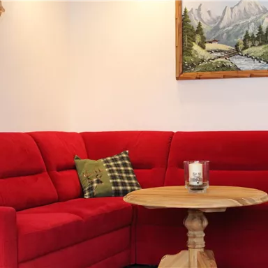 Ein gemütliches Wohnzimmer mit einer roten Couch und einem runden Holztisch. An der Wand hängt ein Landschaftsgemälde.