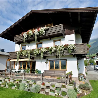 A charming house with flower boxes and a large garden. In the background, gentle hills and a clear sky are visible.