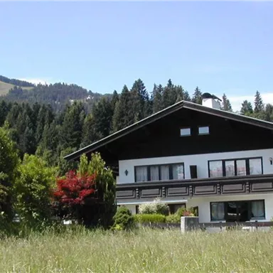 Ein großes Haus umgeben von grünen Wiesen und Bäumen. Im Hintergrund sind sanfte Hügel und ein blauer Himmel sichtbar.