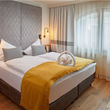 Ein modernes Schlafzimmer mit zwei Betten und einer warmen, gelben Tagesdecke. Große Fenster sorgen für viel Tageslicht und eine einladende Atmosphäre.