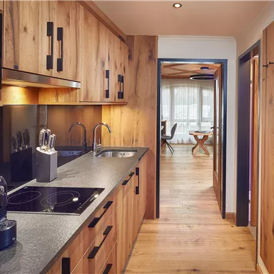 Eine moderne Küche mit Holzmöbeln und einer Kochplatte. Der Blick führt in einen hellen Wohnraum mit Holzboden.