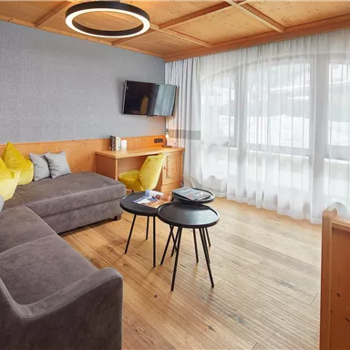 Eine moderne Wohnstube mit einem gemütlichen Sofa und bunten Kissen. Große Fenster lassen viel Licht herein und bieten einen schönen Ausblick.