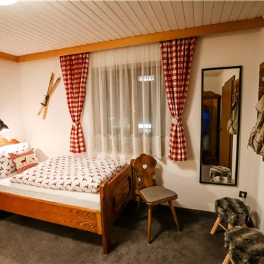 Ein gemütliches Schlafzimmer mit einem Holzbett und karierten Vorhängen. Das Zimmer ist im rustikalen Stil eingerichtet und hat einen Spiegel sowie zwei Stühle.