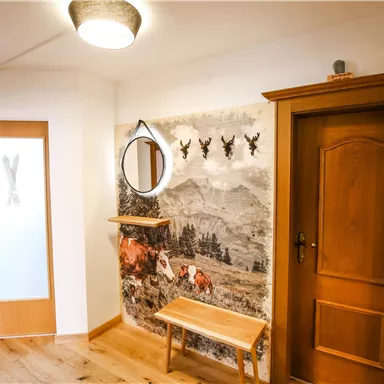 Ein einladender Eingangsbereich mit Holzmöbeln und einem runden Spiegel. An der Wand befindet sich ein dekoratives Alpenmotiv.