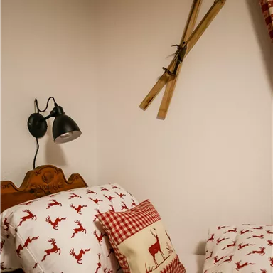 Ein gemütliches Schlafzimmer mit roten Rentier-Mustern auf den Kissen. An der Wand hängen alte Skier und eine Deckenleuchte ist installiert.