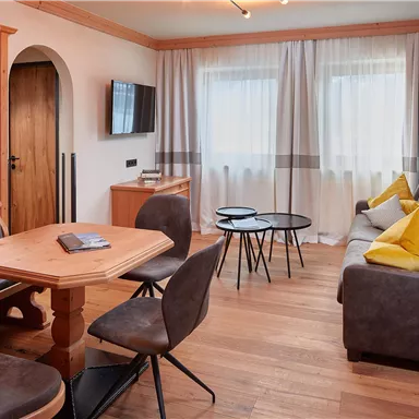 Ein gemütlicher Wohnraum mit einem Holztisch und Stühlen. Die Einrichtung ist modern mit einem grauen Sofa und bunten Kissen.