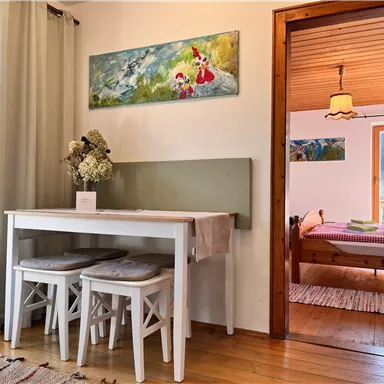 Ein gemütlicher Essbereich mit einem weißen Tisch und Stühlen, dekoriert mit einer Vase. Im Hintergrund ist ein Schlafzimmer mit einem bunten Bett und Fenstern, die auf die Natur blicken.