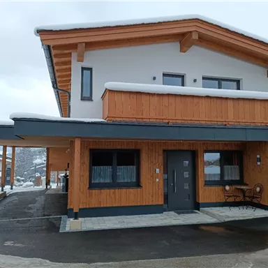 A modern wooden house with a large balcony. It is surrounded by snow and has an inviting facade.