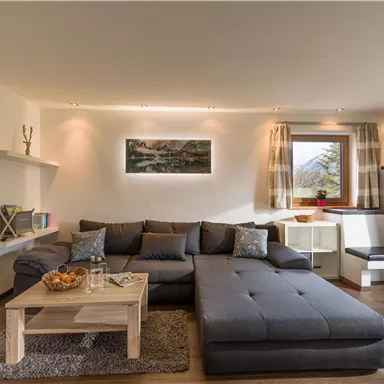 Ein gemütliches Wohnzimmer mit einem großen grauen Sofa und einem Holzkaffee-Tisch. Durch das Fenster scheint Tageslicht, und es gibt Pflanzen und eine Dekoration an den Wänden.