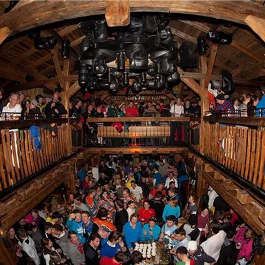 Ein lebhaftes Event in einem Holzgebäude mit vielen Menschen, die sich versammeln. Es gibt eine obere und untere Etage, beide sind gut besucht.