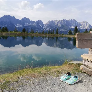 Ein ruhiger See umgeben von Bergen und Bäumen. Neben einem Holzschild stehen ein paar Sportschuhe auf dem Boden.