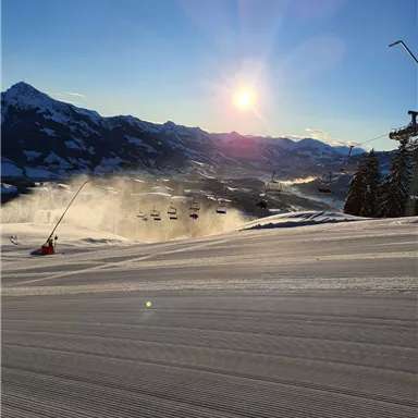 A snowy landscape with freshly groomed slopes and a chairlift. In the background, the sun shines over the mountains.