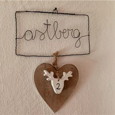 Ein Drahtschild mit der Aufschrift "astberg" hängt an der Wand. Darunter befindet sich ein hölzernes Herz mit einer Zahl und einem Geweih.