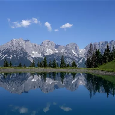 Ein klarer See mit einer ruhigen Wasseroberfläche, die die majestätischen Berge und den blauen Himmel reflektiert. Umgeben von grünen Bäumen und einer idyllischen Landschaft.
