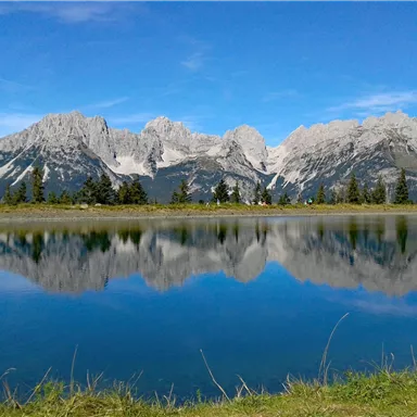 A breathtaking mountain landscape is reflected in the calm waters of a lake. The clear sky and the green meadows draw the viewer into nature.