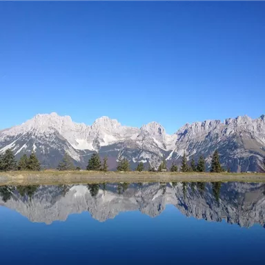 Ein klarer See in den Bergen spiegelt die beeindruckenden Gipfel wider. Der Himmel ist blau und wolkenlos.