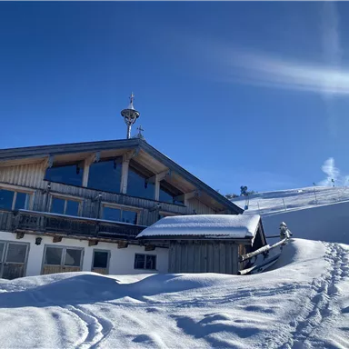 A cozy wooden house in the snow under a clear blue sky. The surroundings are covered with a thick layer of snow.