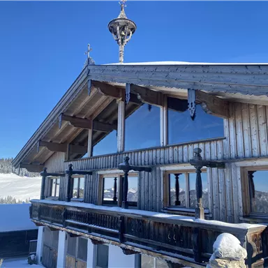 A charming wooden-clad chalet in the snow with large windows and a traditional roof. The hilly winter landscape in the background conveys a tranquil atmosphere.