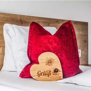 A red pillow on a bed with a wooden heart-shaped sign. The sign reads "Greeting."